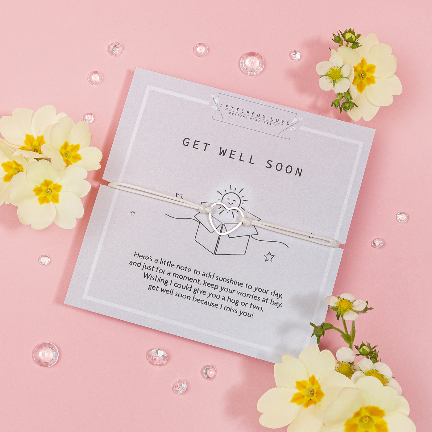 Charming 'Get Well Soon' card on a soft pink background, tied with a delicate white ribbon. The card features a cheerful sun illustration and a comforting message. Beside the card are vibrant yellow flowers with white petals and scattered clear gemstones, evoking a sense of warmth and well-wishes.