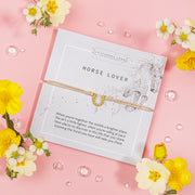 "'HORSE LOVER' gold horseshoe-shaped bracelet on a beige cord, presented on a light grey card with abstract splatter designs. The card features a touching message about the bond between a horse and its rider.
