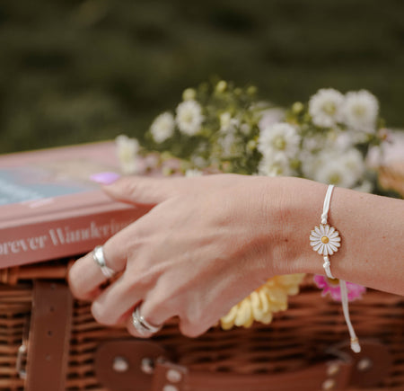 A daisy bracelet on a person's wrist, with a book and flowers in the background.