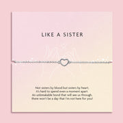 LIKE A SISTER' bracelet featuring a heart charm, presented on a muted pink card with a heartfelt message about the deep bond of friendship that's akin to sisterhood. The card is adorned with delicate pink blossoms and clear crystal gems, set against a soft pink backdrop