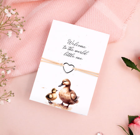 Seeded card featuring a delicate illustration of a mother duck and duckling, with the message 'Welcome to the world little one' in elegant script. A dainty heart-shaped bracelet is attached to the card, tied with natural twine. The card is set against a soft pink background with floral accents, creating a warm and welcoming aesthetic.