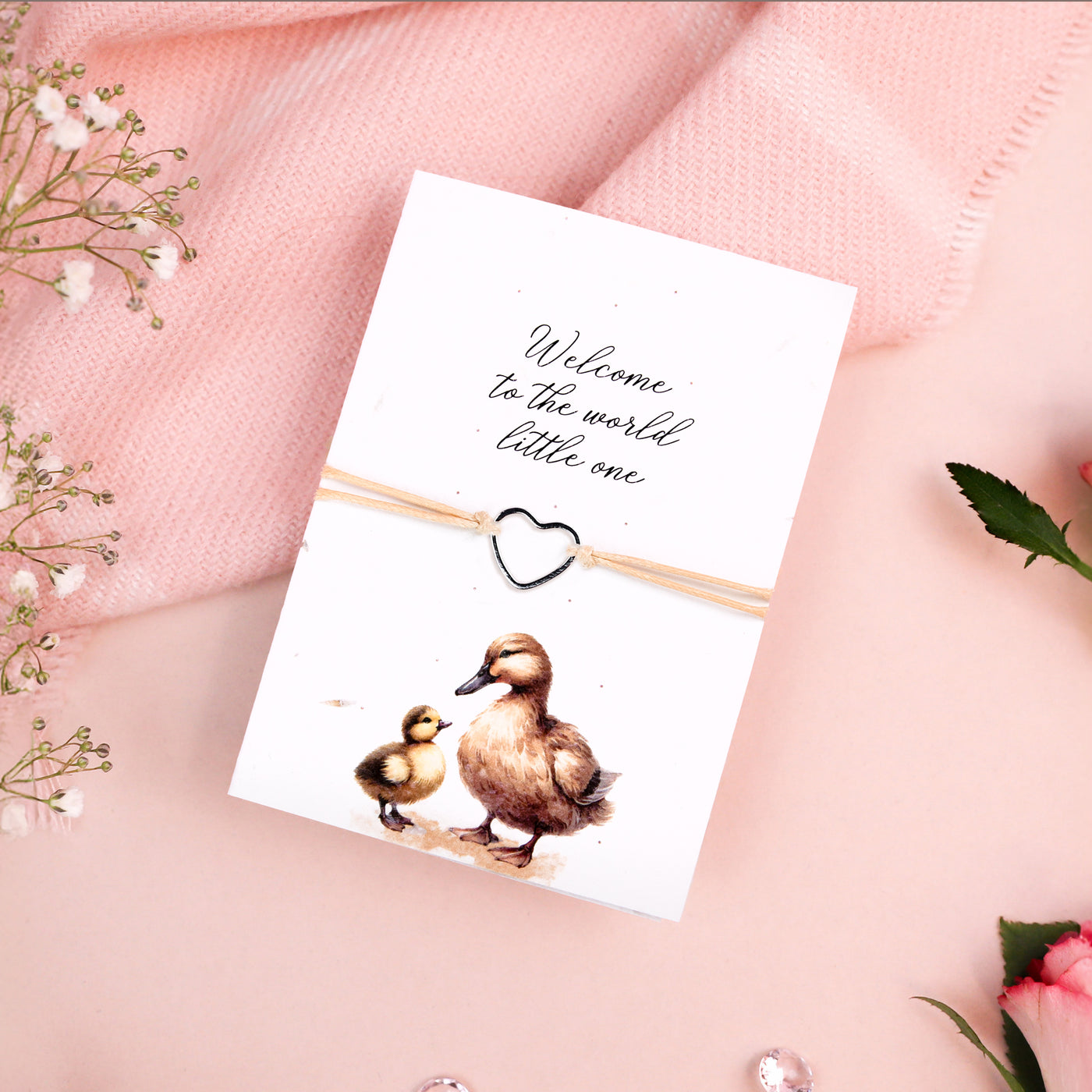 Seeded card featuring a delicate illustration of a mother duck and duckling, with the message 'Welcome to the world little one' in elegant script. A dainty heart-shaped bracelet is attached to the card, tied with natural twine. The card is set against a soft pink background with floral accents, creating a warm and welcoming aesthetic.