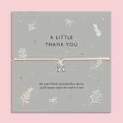 A flat-lay image of a 'A Little Thank You' bracelet gift card. The card is grey with delicate cream botanical and heart illustrations, featuring a thin cream cord bracelet with a small silver bow charm in the center. Below the bracelet, there's a sentimental message: 'My true friend, you're kind as can be, you'll always mean the world to me!' The background is a soft pink.