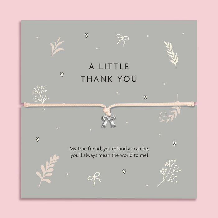 A flat-lay image of a 'A Little Thank You' bracelet gift card. The card is grey with delicate cream botanical and heart illustrations, featuring a thin cream cord bracelet with a small silver bow charm in the center. Below the bracelet, there's a sentimental message: 'My true friend, you're kind as can be, you'll always mean the world to me!' The background is a soft pink.