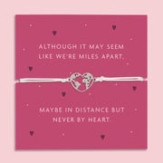 A white adjustable bracelet featuring a heart charm with a world map design, displayed on a deep pink backing card with the message: "Although it may seem like we’re miles apart, maybe in distance but never by heart."