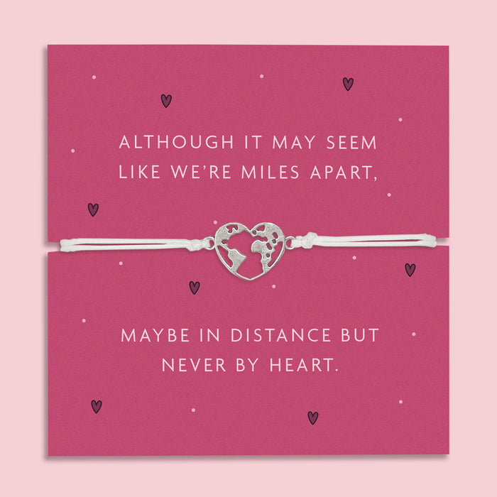 A white adjustable bracelet featuring a heart charm with a world map design, displayed on a deep pink backing card with the message: "Although it may seem like we’re miles apart, maybe in distance but never by heart."