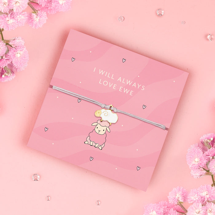 A pink adjustable bracelet featuring an adorable enamel sheep charm, displayed on a pastel pink backing card with the message "I Will Always Love Ewe." Surrounded by soft pink flowers and delicate decorative accents. Perfect for gifting to loved ones.