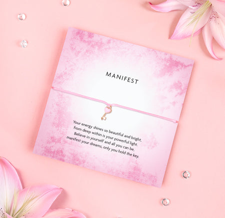 Pink 'Manifest' bracelet with heart charm on a flower-themed greeting card with motivational message, placed on a pink background with pink flowers.