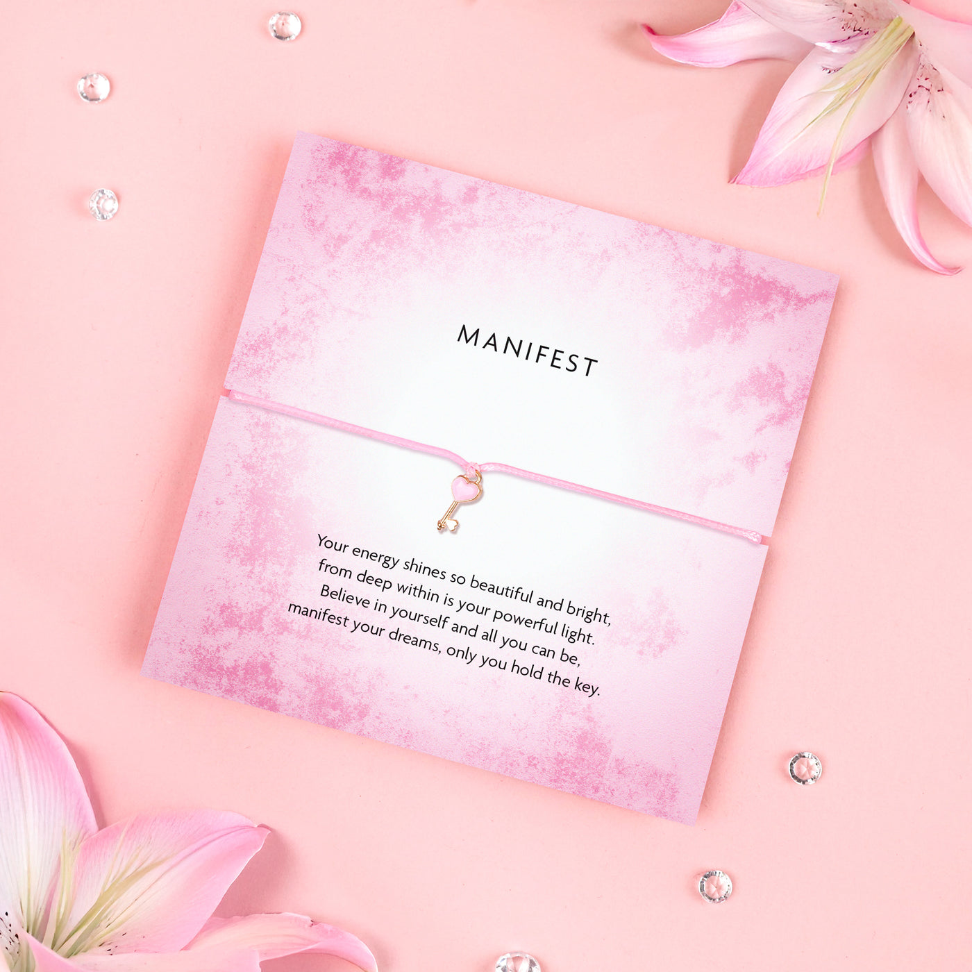 Pink 'Manifest' bracelet with heart charm on a flower-themed greeting card with motivational message, placed on a pink background with pink flowers.