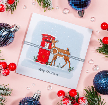 A Christmas greeting card featuring an illustrated deer next to a red postbox amidst falling snow, with a 'Merry Christmas' message below. 