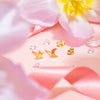 Gold-plated stainless steel star and crescent moon stud earrings with butterfly backs, styled on a soft pink background with floral accents and ribbon.
