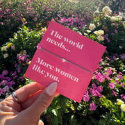 A hand holds a pink folded card with white text reading 'The world needs.