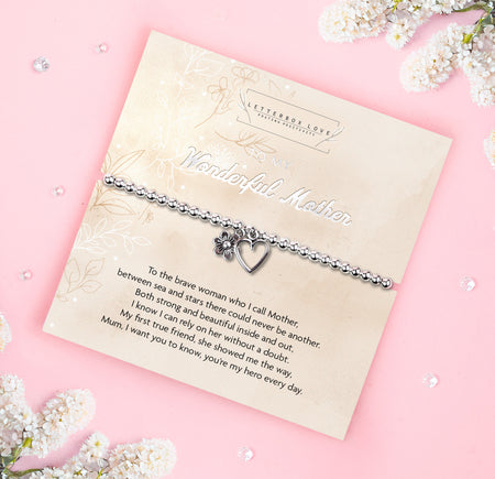 A silver beaded bracelet with a heart and flower charm displayed on a card reading 