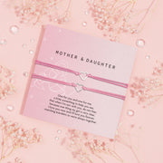 Pair of pink corded bracelets with heart-shaped charms presented on a lavender 'MOTHER & DAUGHTER' card, with a touching message about love and togetherness, adorned with baby's breath flowers and crystal accents on a pastel pink backdrop.