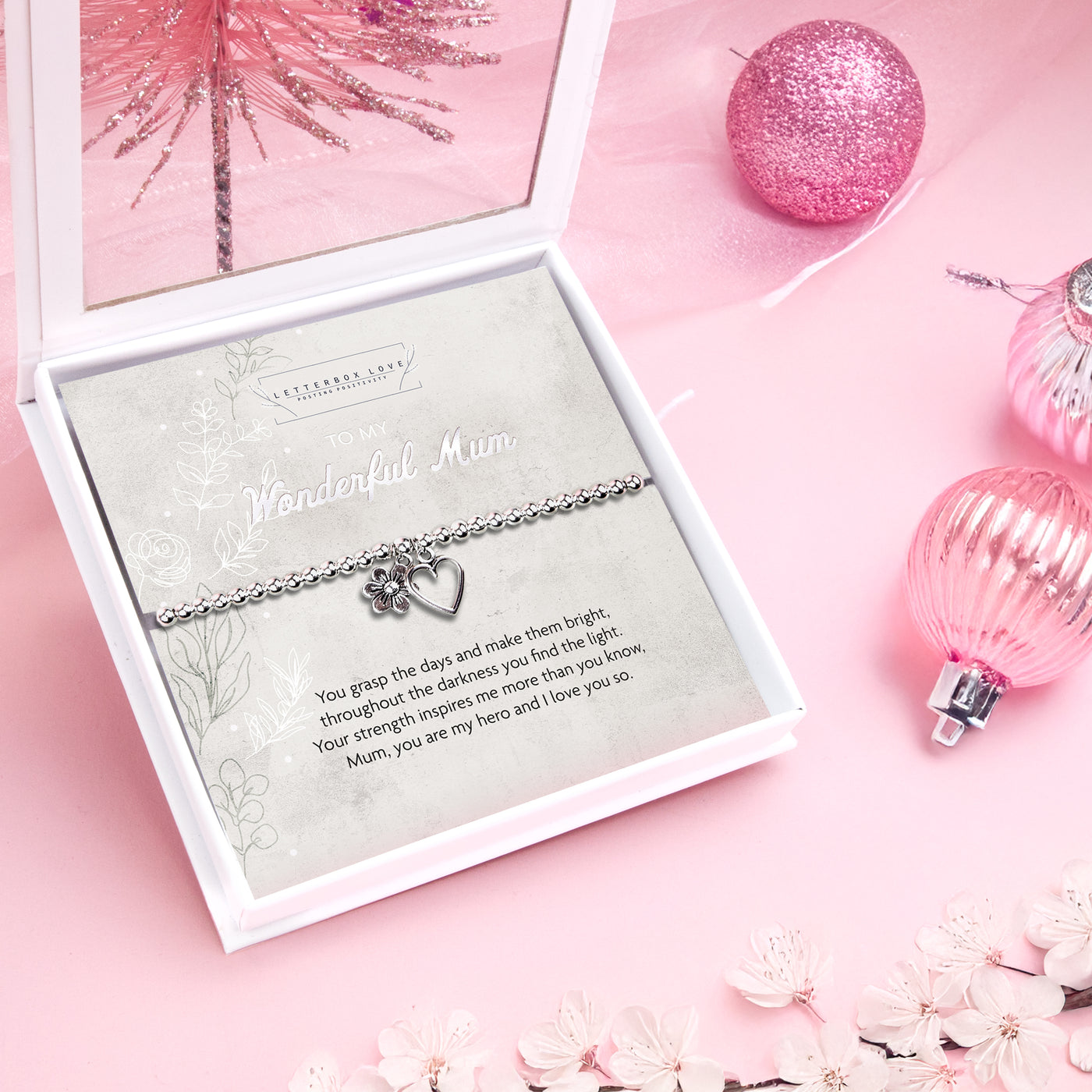 A silver bracelet with a heart and flower charm displayed in a white gift box. The box features a heartfelt message for a mum, set against a soft grey background. Surrounded by pink festive ornaments and floral decorations on a pink surface.