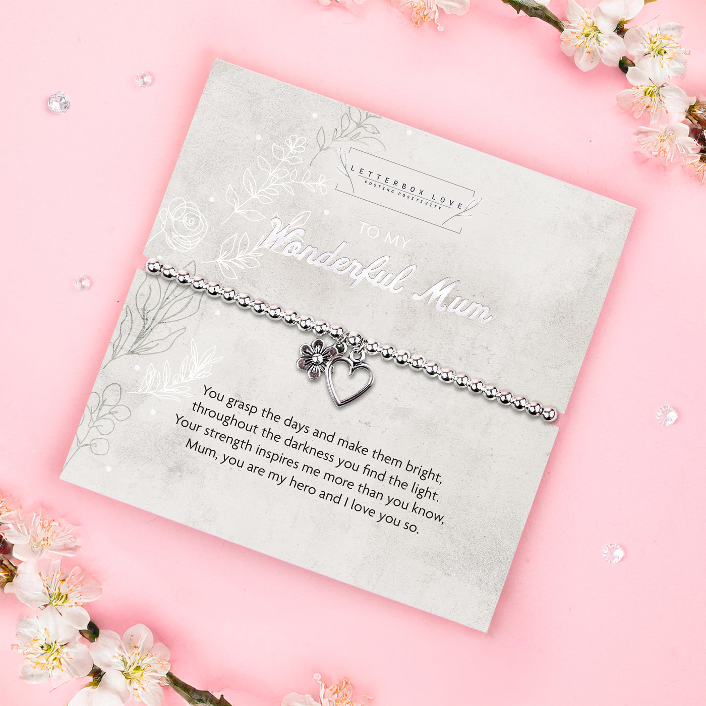 A silver beaded bracelet with a heart and flower charm displayed on a card reading 