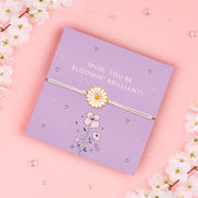 A delicate bracelet with an enamel daisy charm on an adjustable white cord, displayed on a lilac backing card with the message "Mum, You're Bloomin' Brilliant!" Surrounded by soft pink cherry blossoms on a pastel pink background. Perfect for gifting to mums.
