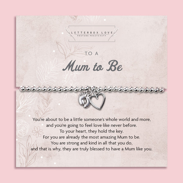 Silver beaded bracelet with heart and baby footprint charms, displayed on a sentimental card reading "To a Mum to Be" with floral details, photographed on a soft pink background with flowers and gems.
