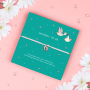 A turquoise card with the words "MUMMY TO BE" in white uppercase letters. The card is decorated with soft pink hearts, stars, and an illustration of a stork carrying a baby wrapped in a bundle. A delicate silver baby feet charm bracelet on a soft ivory cord is wrapped around the card. Below, the message reads: "As your heart grows, your world will too, how blessed they are to have a mummy like you." The background is plain white, highlighting the bracelet and card design.