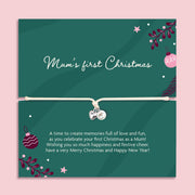 A square poem card with a thin white bracelet across the center and small heart and pearl charms. The card is dark green with white script reading 'Mum's first Christmas' and smaller white text below, decorated with Christmas ornaments, stars, and foliage on a light pink background.