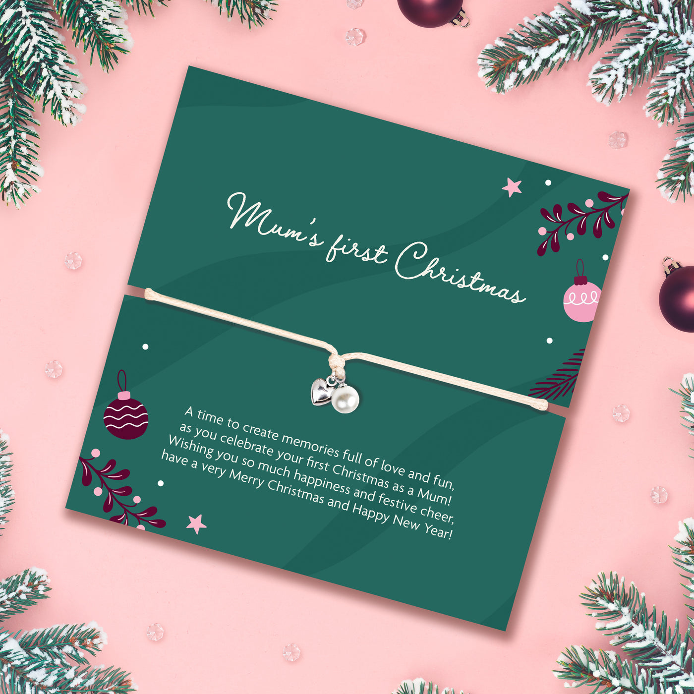 A Christmas-themed card for 'Mum's first Christmas' featuring a delicate white cord bracelet with a silver heart and pearl charm, displayed on a pink background decorated with pine branches and ornaments.