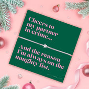 Green card with pink text and a silver bracelet on a pink background with Christmas decorations.