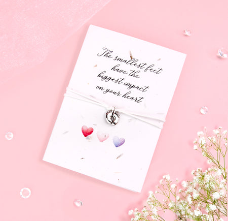 Seeded card with the message 'The smallest feet have the biggest impact on your heart' in elegant script. Attached is a delicate bracelet with a small silver baby feet charm. The card features three watercolor hearts in shades of red, pink, and purple, set against a soft pink background with scattered clear gems and baby's breath flowers for a sweet and gentle touch.