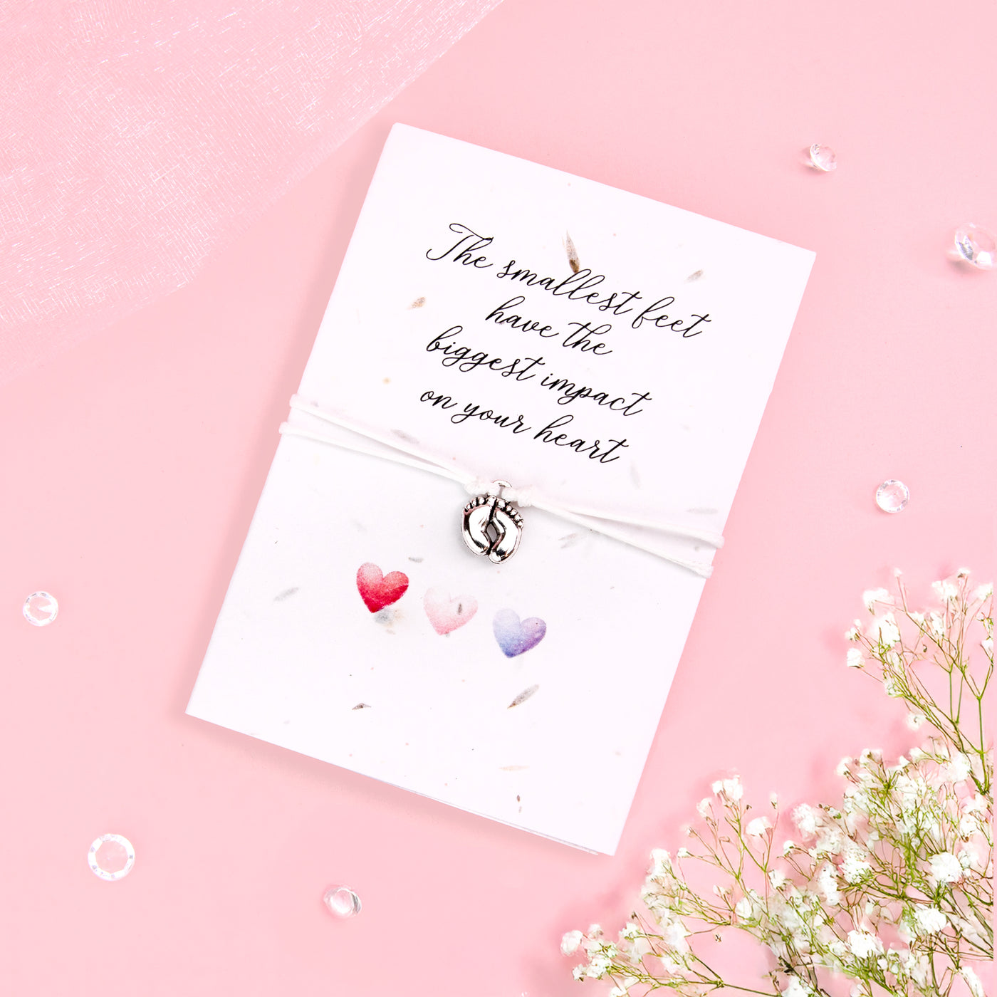 Seeded card with the message 'The smallest feet have the biggest impact on your heart' in elegant script. Attached is a delicate bracelet with a small silver baby feet charm. The card features three watercolor hearts in shades of red, pink, and purple, set against a soft pink background with scattered clear gems and baby's breath flowers for a sweet and gentle touch.