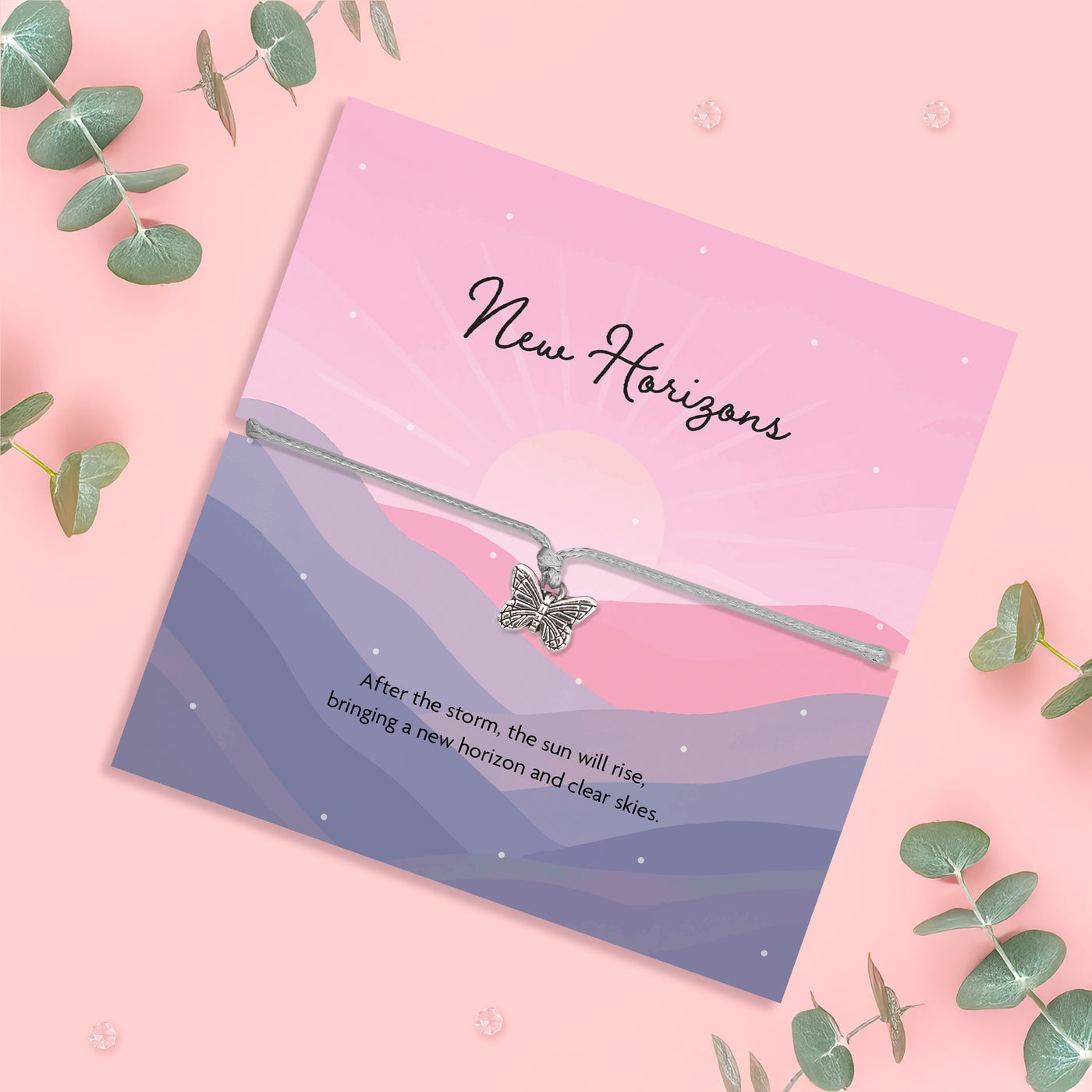 Bracelet with butterfly charm on a scenic card with 'New Horizons' text, surrounded by eucalyptus leaves on a pink background.