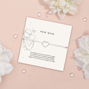 An elegant 'New Mum' bracelet with a dainty heart charm, displayed on a pastel pink background and sparkling crystals, symbolizing the gentle embrace of motherhood.