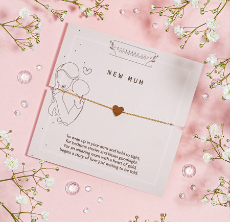 A delicate gold bracelet with a tiny heart charm presented on a pastel pink backdrop with the inscription 'NEW MUM' and a touching poem about motherhood. 