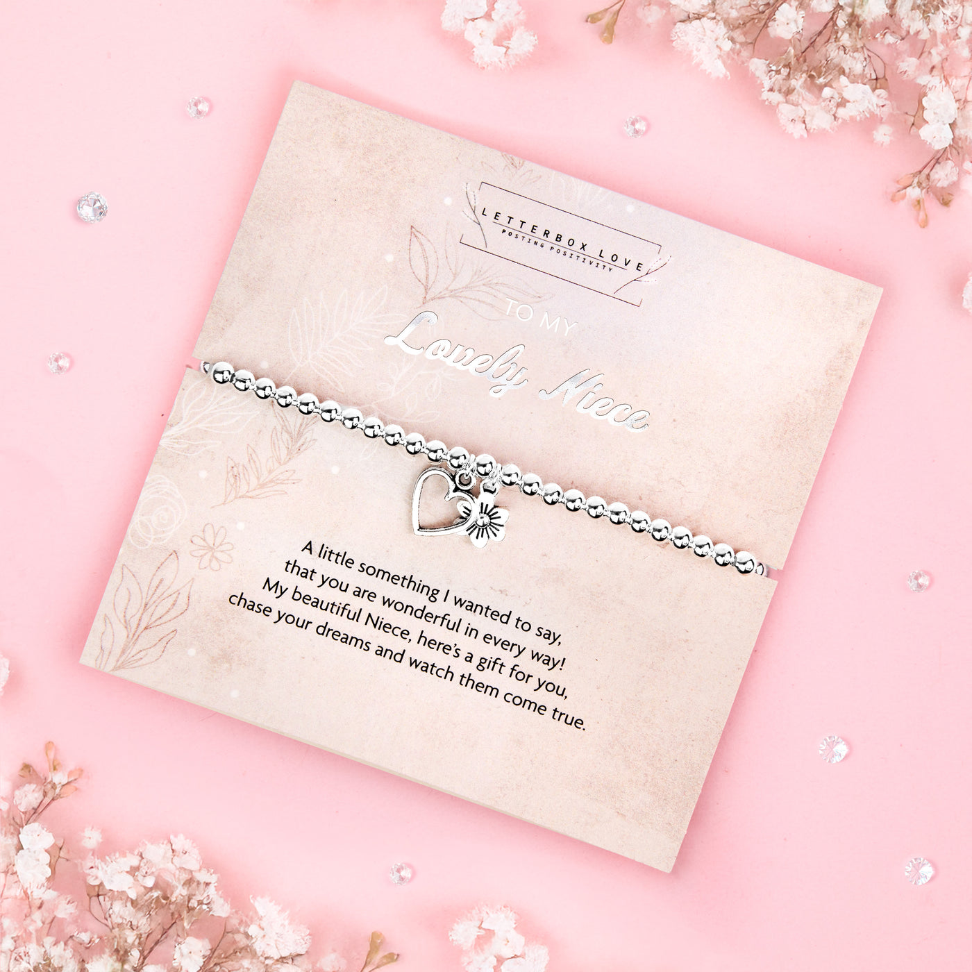 A silver beaded bracelet with a heart and flower charm displayed on a card reading 