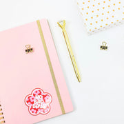 Pink notebook with a gold pen and decorative clip on a white background with a 'no bad days' girlie sticker