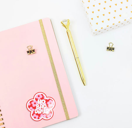 Pink notebook with a gold pen and decorative clip on a white background with a 'no bad days' girlie sticker