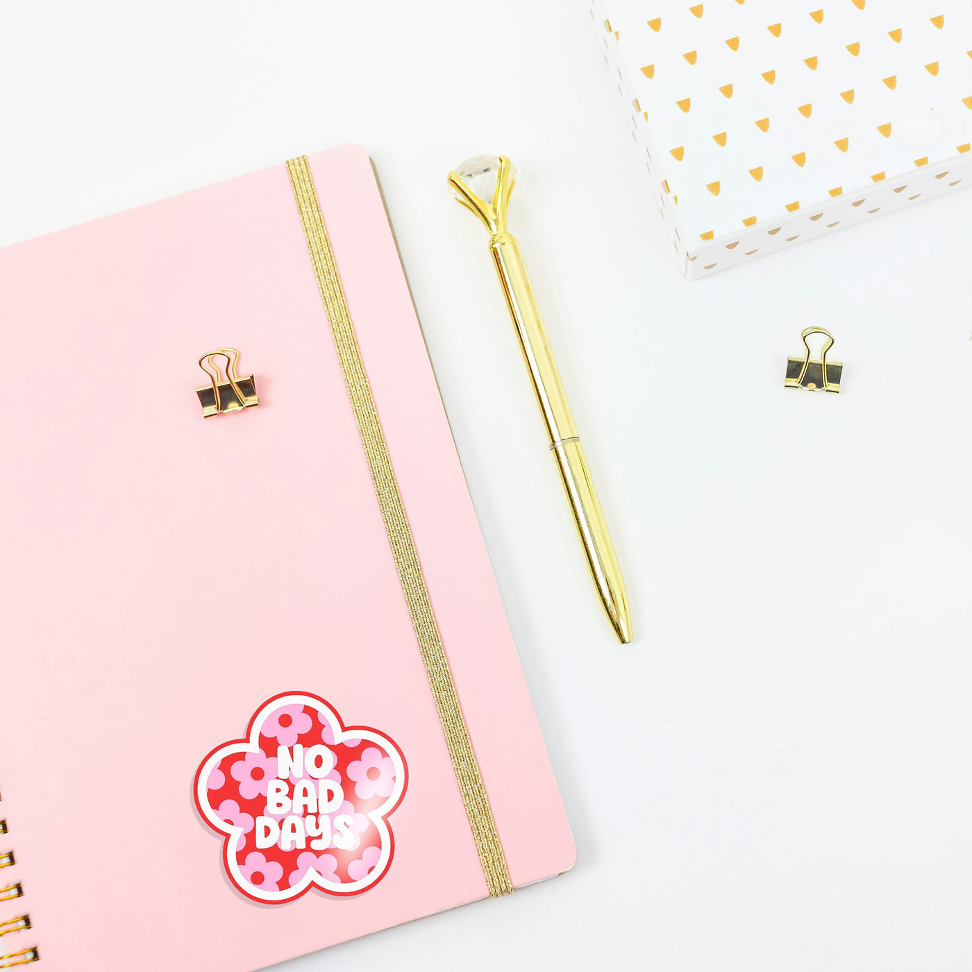 Pink notebook with a gold pen and decorative clip on a white background with a 'no bad days' girlie sticker