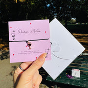 A hand holds a pink card and white envelope featuring a small wine glass charm on a black bracelet, photographed outdoors in a park with trees and a bench in the background.