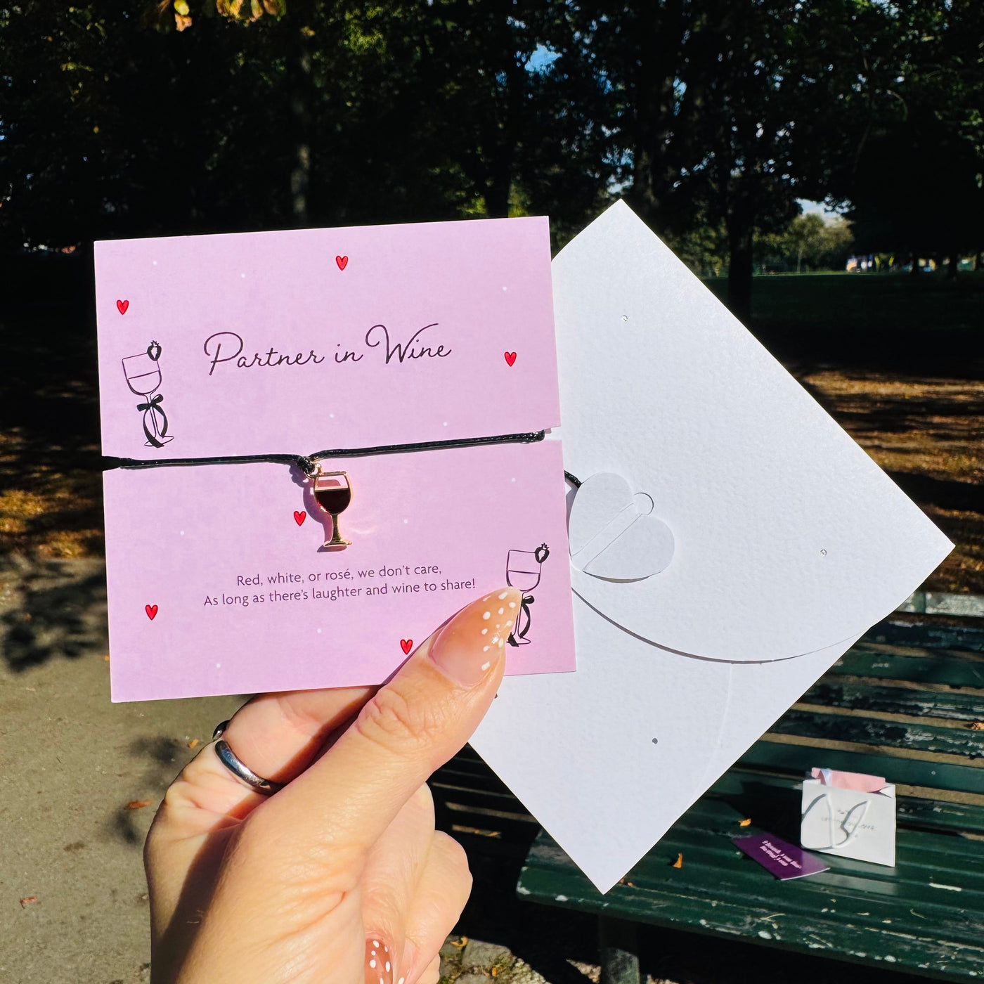 A hand holds a pink card and white envelope featuring a small wine glass charm on a black bracelet, photographed outdoors in a park with trees and a bench in the background.