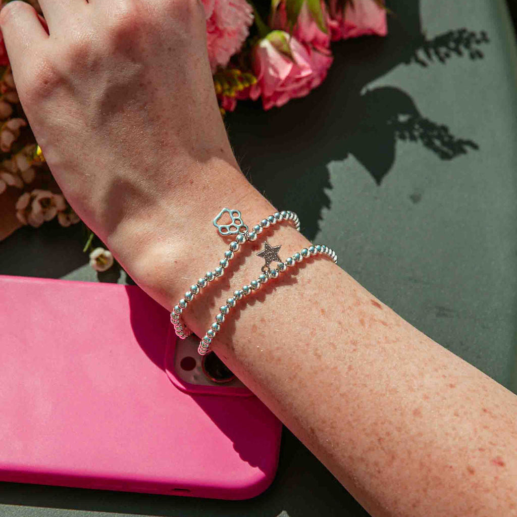 Woman's arm with silver-plated bracelets on a pink phone case and flowers in the background. The bracelets have a dog paw charm and a star.