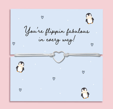 Flippin’ Fabulous Penguin Bracelet displayed on a light blue festive card decorated with cartoon penguins, tiny hearts, and falling snow dots. The bracelet features a silver-tone open heart charm on a grey adjustable cord, making it a fun and meaningful Christmas jewellery gift. Perfect as a Secret Santa present, stocking filler, or festive gift for penguin and animal lovers. 