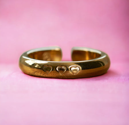 Gold stainless steel adjustable ring with three embossed heart designs, displayed against a vibrant pink background.