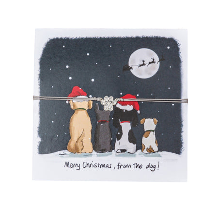 Festive greeting card featuring an illustration of four dogs in Santa hats looking up at a night sky with snowflakes, a full moon, and Santa's sleigh flying past. A paw-shaped bracelet charm with glittering stones is tied to the card with a string, enhancing the festive appearance. Below the scene is the humorous phrase 'Merry Christmas, from the dog!' 