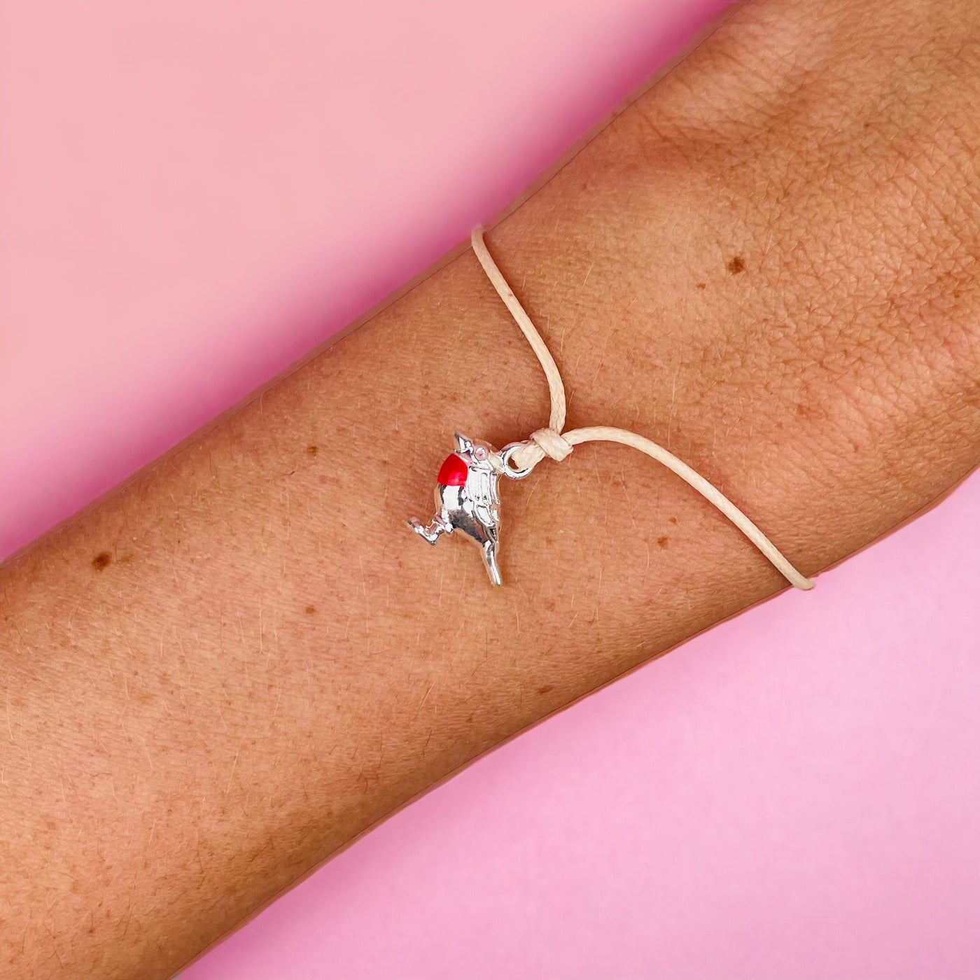 A delicate bracelet with a single, intricately crafted robin charm, featuring a red breast, perched on a soft, natural beige cord against a person's wrist.