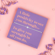 A lavender-colored bracelet card titled "Positivity" from Letterbox Love, featuring a small star charm on a grey string bracelet. The card includes the message: "A little positivity to see you through... To give you strength in all you do." The setting is a soft pink background with small purple flowers and decorative gems scattered around.