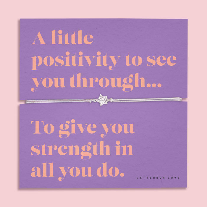 A lavender-colored bracelet card titled "Positivity" from Letterbox Love, featuring a small star charm on a grey string bracelet. The card includes the message: "A little positivity to see you through... To give you strength in all you do." The setting is a soft pink background with small purple flowers and decorative gems scattered around.