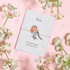 The Little Robin - Seeded  A seed paper card with a watercolor illustration of a robin and a red heart charm on a brown bracelet, set against a pink background. The card reads 