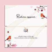 A Christmas bracelet set featuring a top card with the script text 'Robins appear' to bring comfort in the festive season. The bracelet is adjustable with a small silver robin charm on a cream cord, set against a solid pink background.