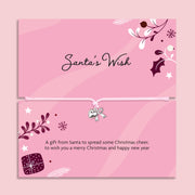 A pink Christmas-themed card or gift card set with decorative illustrations, the title 'Santa's Wish' on the top card, and a small silver bow charm tied across the bottom card. The design includes holly, pine branches, stars, and a gift box, all on a soft pink background. A perfect gift for Secret Santa.
