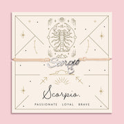 Scorpio zodiac bracelet with silver calligraphy-style “Scorpio” charm and dangling heart on a beige cord, displayed on a celestial cream and gold card with traits: Passionate, Loyal, Brave.

