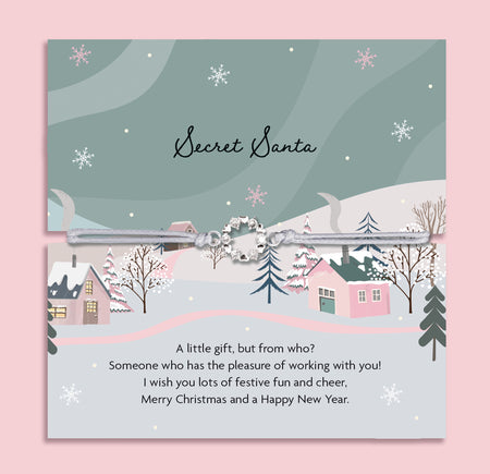 Secret Santa card with a winter village scene and festive text on a pink background, with a beautiful christmas wreath on a friendship bracelet