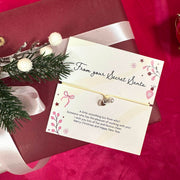 A small silver heart & pearl charm attached to a cream card that reads 'From your Secret Santa,' styled on top of a dark red gift box with a white ribbon and holiday greenery on a red fabric background.